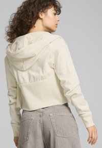 Felpa corta color crema con cintura e retro a coste texture. Dotata di cappuccio con cordini e maniche lunghe. Pantaloni grigi visibili sotto.