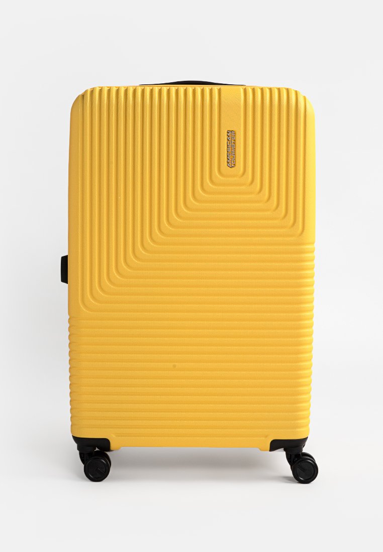 Valigia rigida gialla con scanalature verticali, chiusura con zip e quattro ruote nere per la mobilità. Include un logo del marchio sul davanti.