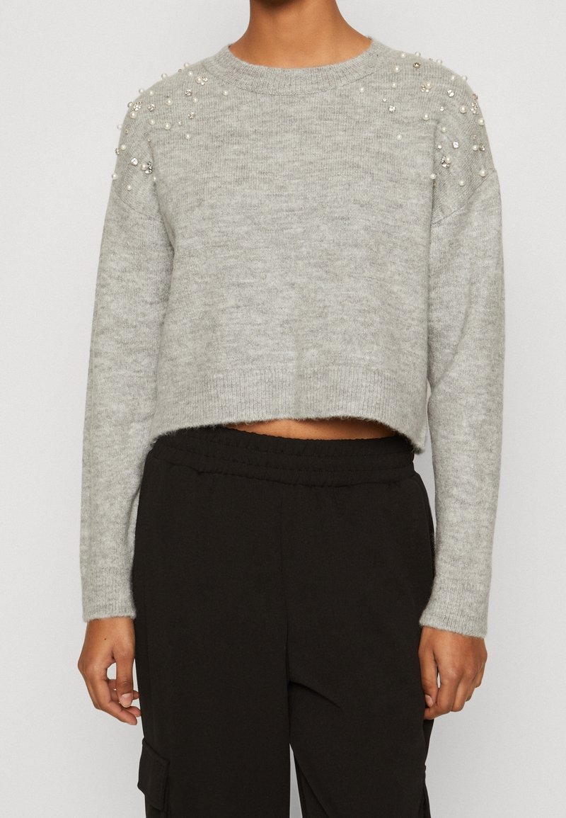 Pull gris court avec décorations en perles et strass sur les épaules, porté avec un pantalon noir taille haute.