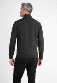 Dunkelgrüner Pullover mit hohem Kragen, gerippter Textur und langen Ärmeln. Gemustertes Gewebe und figurbetontes Design. Kombiniert mit schwarzer Hose.