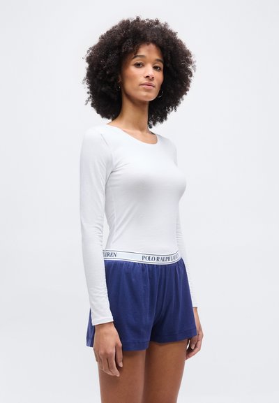 Γυναίκα με σγουρά μαλλιά που φοράει λευκό μακρυμάνικο πουκάμισο και σκούρο μπλε σορτς Polo Ralph Lauren, στέκεται μπροστά σε απλό ανοιχτό φόντο.