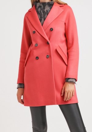 Femme portant un manteau corail à double boutonnage avec des boutons foncés par-dessus un pull gris à motif et un pantalon en cuir noir.