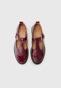 Burgundowe lakierowane buty typu Mary Jane z okrągłym noskiem, wycięciami po bokach, paskiem z klamrą oraz czarną gumową podeszwą.
