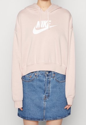 Person trägt einen hellrosa Nike-Hoodie mit weißem Logo und blauen Jeansrock, steht vor einfarbigem Hintergrund. Oberkörper sichtbar.