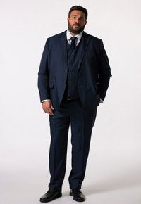 Dunkelblauer Anzug mit einem Zweiknopfjackett, passender Weste und schmal zulaufenden Hosen. Getragen mit einem weißen Hemd und einer dunklen Krawatte. Schwarze Dressschuhe.