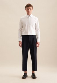 Witte overhemdd met een puntkraag, gecombineerd met marineblauwe, geplooide broek en zwarte leren schoenen. Effen achtergrond.