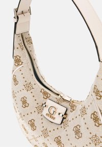 Sac à main beige Guess avec motif monogramme marron, fermeture éclair dorée, et plaque logo dorée "G Guess" sur une surface en tissu texturé.