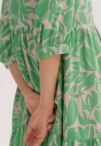 Ręce splecione za plecami, ubrana w luźną, pastelową różową sukienkę z jasnorudym zielonym wzorem liści i rękawami o długości trzech czwartych z guzikowymi mankietami.