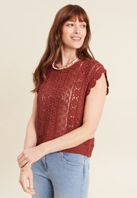 Top all'uncinetto corto rosso ruggine con bordi scallop, design testurizzato e orlo a coste. Indossato sopra jeans azzurri chiari, con una collana di perle.