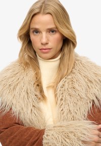 Beige faux fur krave med lange, teksturerede strå over en rustrød cordjakke. Lagdelt ribbet cremefarvet turtleneck under kraven.