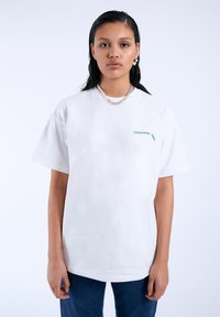 Bílé bavlněné tričko s krátkým rukávem, s zeleným nápisem "SLEEP GOOD TIMES" na levé straně. Nosené s modrými džíny.