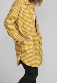 Hnědo-žlutá kostkovaná flanelová košile s knoflíkovým zapínáním, límcem, bočními kapsami a uvolněným střihem. Obsahuje dlouhé rukávy a zakřivený lem.