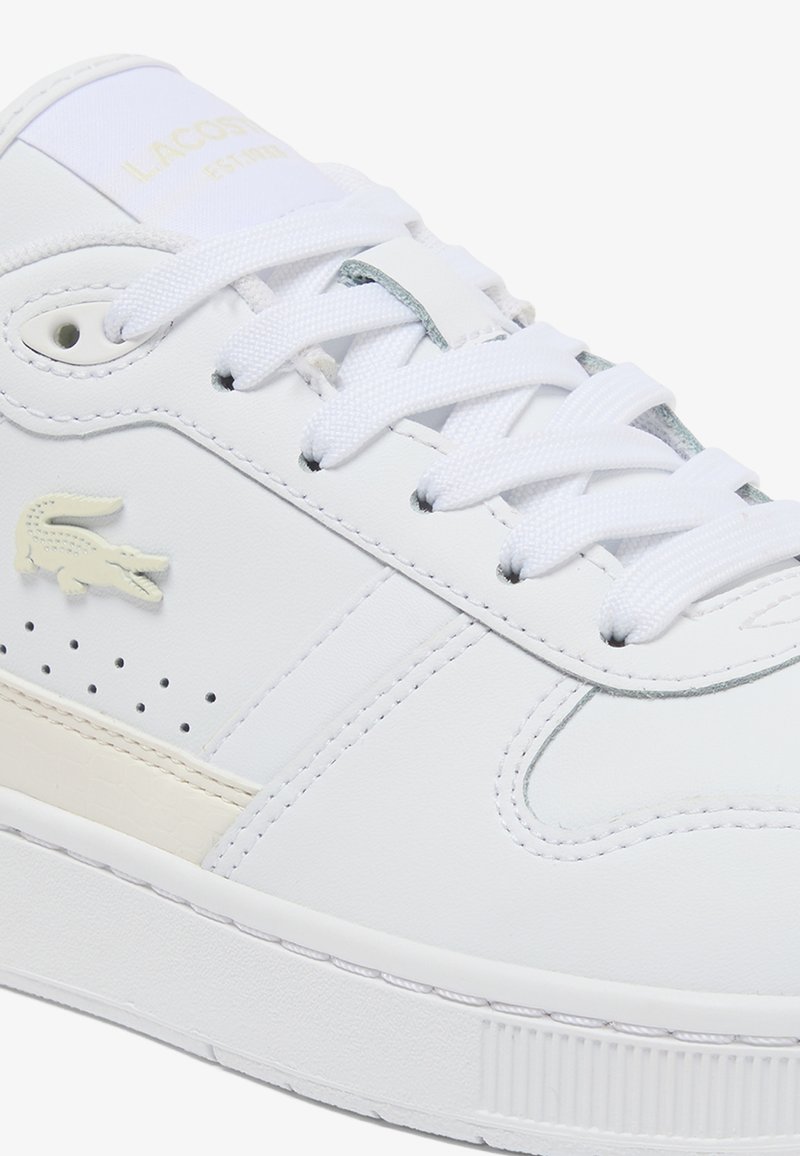 White Lacoste low-top sneaker with white laces, perforated side vents, beige logo patch, and textured rubber sole.