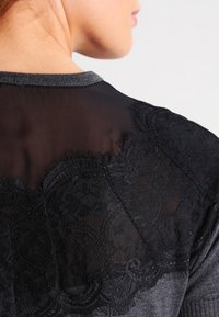 Accenti in pizzo nero sovrappongono un pannello posteriore trasparente, corredato da un tessuto grigio texturizzato e un collo a coste scuro.