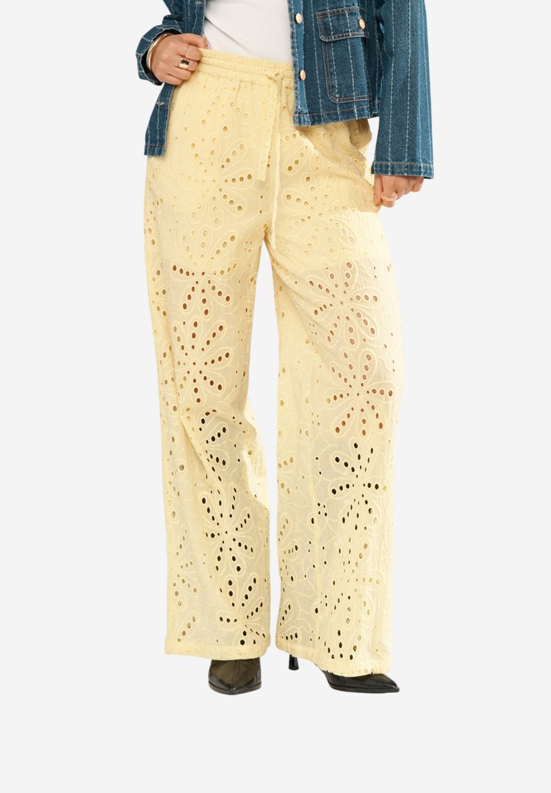 Pantalon jaune à jambes larges avec broderie florale ajourée, porté avec des talons noirs pointus et une veste en jean par-dessus un haut blanc.