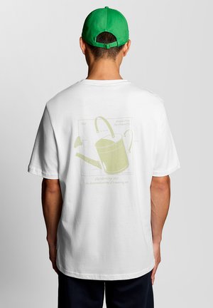 Mann trägt weißes T-Shirt mit gelber Gießkanne als Illustration und grüne Kappe, steht mit dem Rücken zur Kamera vor einem einfarbigen Hintergrund.
