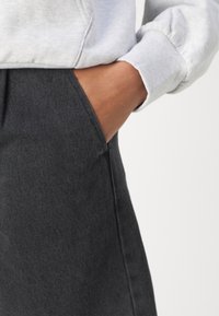 Pravá ruka zasunutá do kapsy tmavě šedých kalhot, oblečená v světle šedé mikině s žebrovaným manžetem, na bílém pozadí.