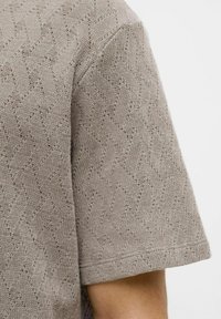 Vaaleanharmaa neulottu lyhythihainen paita, joka esittelee sahalaitakuviota ja teksturoitua kangasta. Hihansuut on viimeistelty siististi.