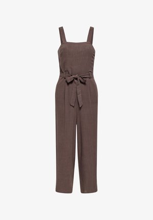 Femme debout, les bras croisés, portant une combinaison marron sans manches à jambes larges, ceinture nouée à la taille et chaussures plates noires sur fond uni.