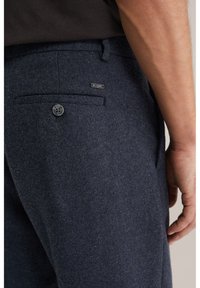 Prim-plan al buzunarului pantalonilor din lână albastru închis ai unui bărbat, cu un nasture și o etichetă mică a brandului, alături de o parte din brațul și mâna sa.