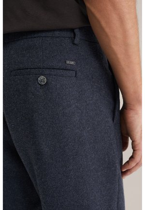 Zbliżenie na kieszeń męskich ciemnoniebieskich wełnianych spodni z guzikiem i małą metką z logo, obok fragmentu jego ramienia i dłoni.