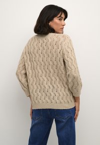 Maglione lavorato a maglia beige con un motivo a onde, dotato di maniche ampie e orlo a costine. Indossato con jeans in denim blu, che mostrano una vestibilità rilassata.