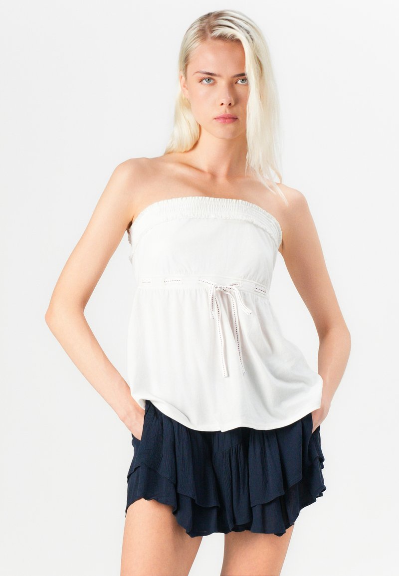 Weißes trägerloses Top mit gerafftem Oberteil und vorderem Bindeband, kombiniert mit einer dunkelblauen, rüschenverzierten Shorts. Glatter Stoff, lockere Passform und lässiges Design.