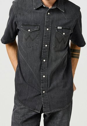 Mann trägt ein kurzärmeliges schwarzes Jeanshemd mit Druckknöpfen und zwei vorderen Klappentaschen, kombiniert mit schwarzen Jeanshosen.