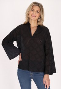 Schwarzes Tunika-Top mit V-Ausschnitt, das über rautenförmige Ausschnittmuster, weite Glockenärmel und einen leichten Stoff verfügt. Kombiniert mit blauen Jeans.