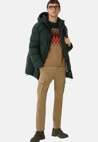 Man wearing green puffer jacket over patterned brown sweater, khaki cargo pants, and dark hiking shoes, standing with one hand in pocket.