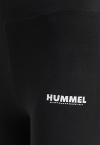 Czarne legginsy sportowe wykonane z elastycznej tkaniny. Zawierają białe logo z napisem „HUMMEL” oraz „SPORTSWEAR SINCE 1923” przy dolnej krawędzi.