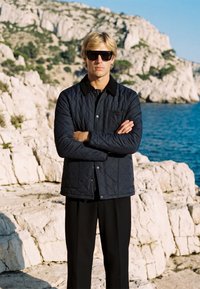 Homme blond portant une veste matelassée noire et des lunettes de soleil, debout, les bras croisés, sur une côte rocheuse avec la mer bleue et des falaises en arrière-plan.