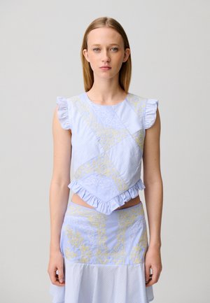 Femme aux cheveux blonds raides portant un haut sans manches bleu clair et une jupe assortie avec des motifs ornementaux jaunes et des détails à volants.