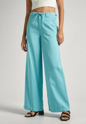 Pantalones de pierna ancha turquesa hechos de una tela ligera, con cintura elástica, bolsillos laterales y dobladillos sin terminar. Combinados con sandalias negras.