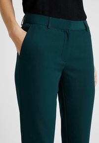 Byxor i djupt teal, tillverkade av mjukt tyg. Har en platt front med snedställda fickor och en skräddarsydd passform. Slät textur och strukturerad design.