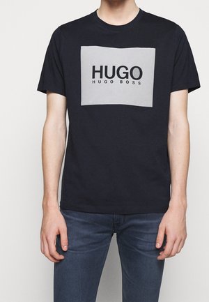 Osoba nosząca czarny T-shirt z dużym szarym prostokątem i tekstem "HUGO HUGO BOSS" nadrukowanym z przodu, w połączeniu z niebieskimi dżinsami.