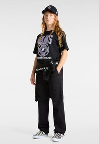 Černé grafické tričko s fialovým textem, černé kalhoty a černobílé kostkované slip-on boty. Stylizováno s černou kšiltovkou a svetrem uvázaným v pase.