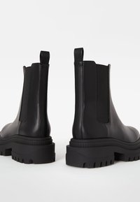 Černé kožené kotníkové boty s elastickými bočními panely, silnou gumovou podrážkou a táhly na patě. Design má robustní, texturovanou podrážku.