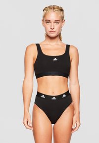 Crni sportski grudnjak i visoke gaćice s širokim elastičnim pojasom. Na prednjoj strani se nalazi bijeli Adidas logo. Elegantan, minimalistički dizajn.