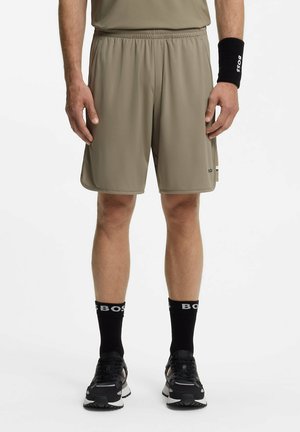 Man draagt khaki sportshorts, een zwart BOSS polsbandje, zwarte BOSS sportsokken en zwarte sportschoenen, staand tegen een effen achtergrond.