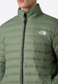 Giacca a vento verde con colletto alto, dotata di cuciture orizzontali, zip frontale e logo bianco sul lato sinistro del petto.