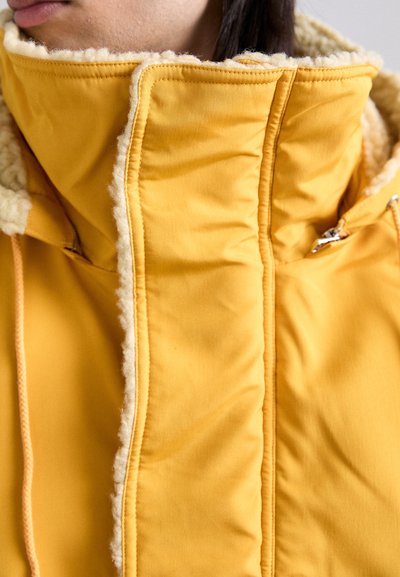 Yellow padded jacket featuring a high collar with soft fleece lining, visible stitching, and a zippered pocket on the side.