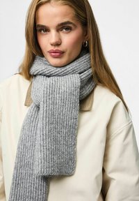 Écharpe en tricot côtelé gris portée sur un manteau beige clair. L'écharpe a une texture douce et tombe de manière lâche, avec une extrémité légèrement plus large.