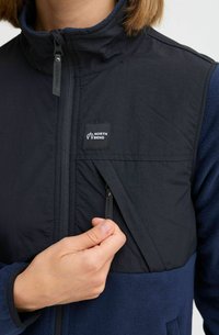 Veste hybride noire et marine avec une poche poitrine zippée, dotée d'un col haut et d'un tissu texturé. Logo "North Bend" sur la poche.