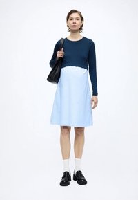 Μπλε ναυτικό τοπ με φαρδύ, ριγέ φούστα σε ανοιχτό γαλάζιο. Η σιλουέτα είναι χαλαρή. Φορεμένο με μαύρα μοκασίνια και λευκές κάλτσες, και μαύρη τσάντα.