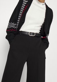 Černý cardigan s červenými a bílými pruhy, nošený přes bílý žebrovaný top, spárovaný s černými kalhotami s vysokým pasem a černým opaskem.
