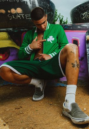 Chaqueta verde con cremallera y detalles en blanco, logo de trébol, pantalones cortos verdes, zapatos gris claro sin cordones y calcetines blancos. Sentado/a contra un fondo colorido.