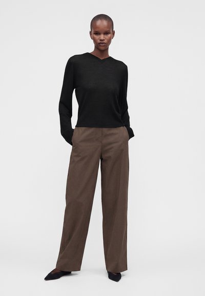 Femme debout portant un haut noir à manches longues, un pantalon marron ample et des chaussures pointues noires sur un fond clair uni.