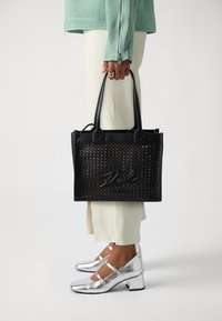 Juoda nertinė rankinė su odiniais akcentais ir iškilmingu logotipu, pasižyminti tvirtais rankenėlėmis. Dėvima su kreminės spalvos kelnėmis ir sidabrinėmis aukštakulnėmis bateliais.