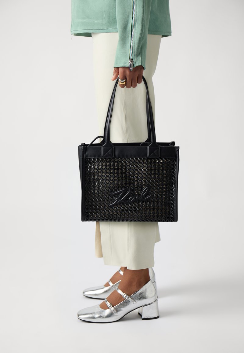 Juoda nertinė rankinė su odiniais akcentais ir iškilmingu logotipu, pasižyminti tvirtais rankenėlėmis. Dėvima su kreminės spalvos kelnėmis ir sidabrinėmis aukštakulnėmis bateliais.